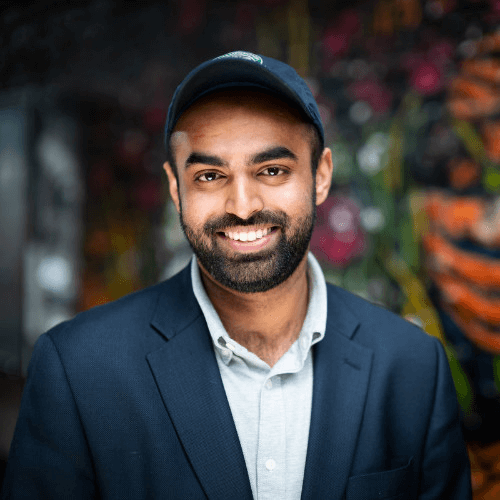 Varun Manoj, Senior Solutions Engineer at Snyk, wearing a navy blue suit jacket with a white collar shirt and a navy blue cap.