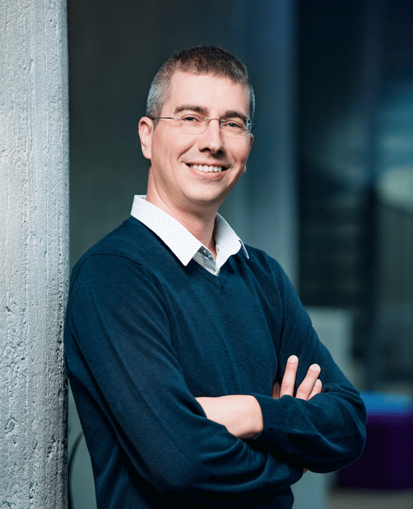 An image of Jani Ruuskanen in a navy blue shirt smiling for camera while leaning to a wall and crossing his arms.